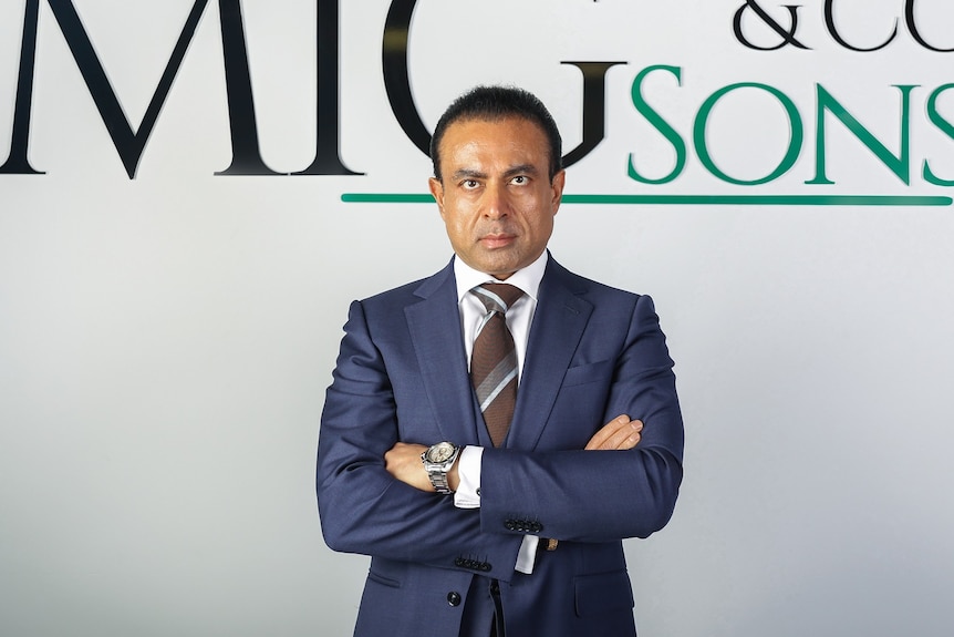 A man in a suit standing with his arms crossed in front of his business logo which reads "MIG Sons & Co"