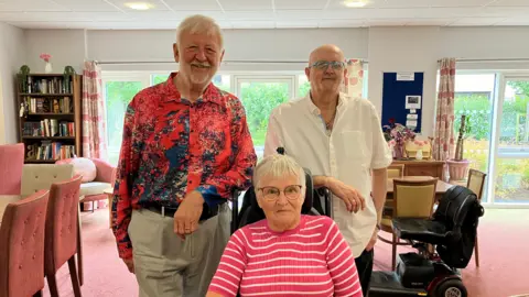 BBC Two men are stood in a lounge with furniture and a mobility scooter behind them. One has a bright red and blue shirt, the other is wearing a white one. There is a woman with a red and white striped top, who is seated in front of them