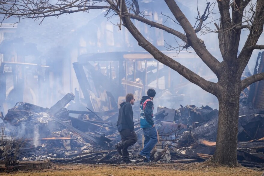 Two people walk past a pile of charred debris of a burned house. Smoke rises from the debris, and the burnt frame of a house is in the background.