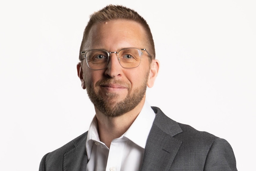 A man in clear frame glasses, a grey suit jacket and white shirt