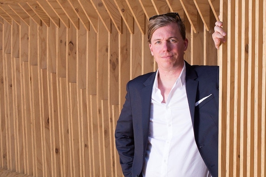 A man with blonde hair and sunglasses standing in front of a wall in a suit jacket